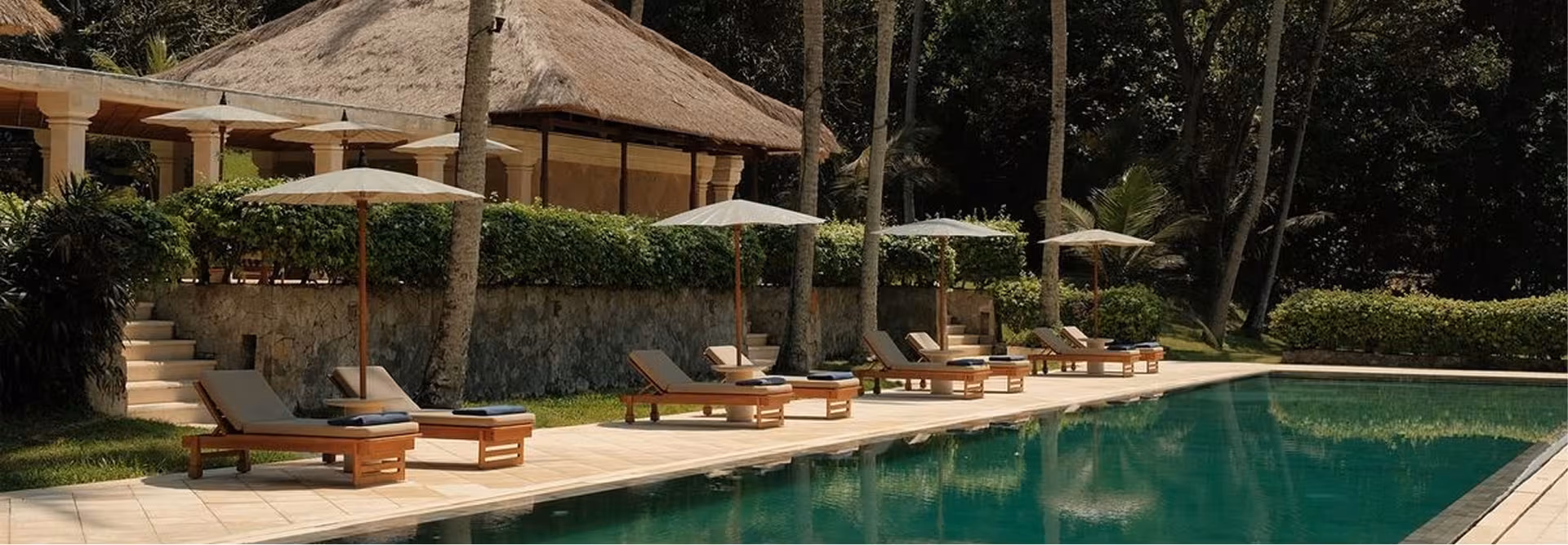 Wooden sun loungers and umbrellas beside a green pool, with a thatched-roof pavilion and palm trees behind